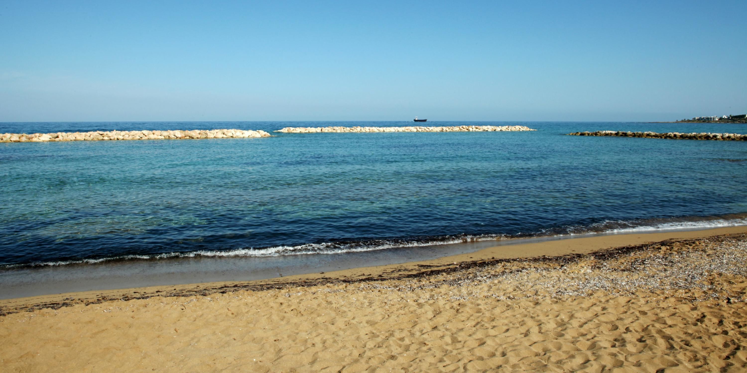 A beach with clear blue water, a rocky breakwater, and a distant boat.