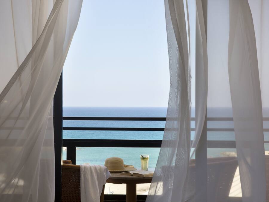 A balcony with a view of the ocean, a table with a hat and a book, and sheer curtains.