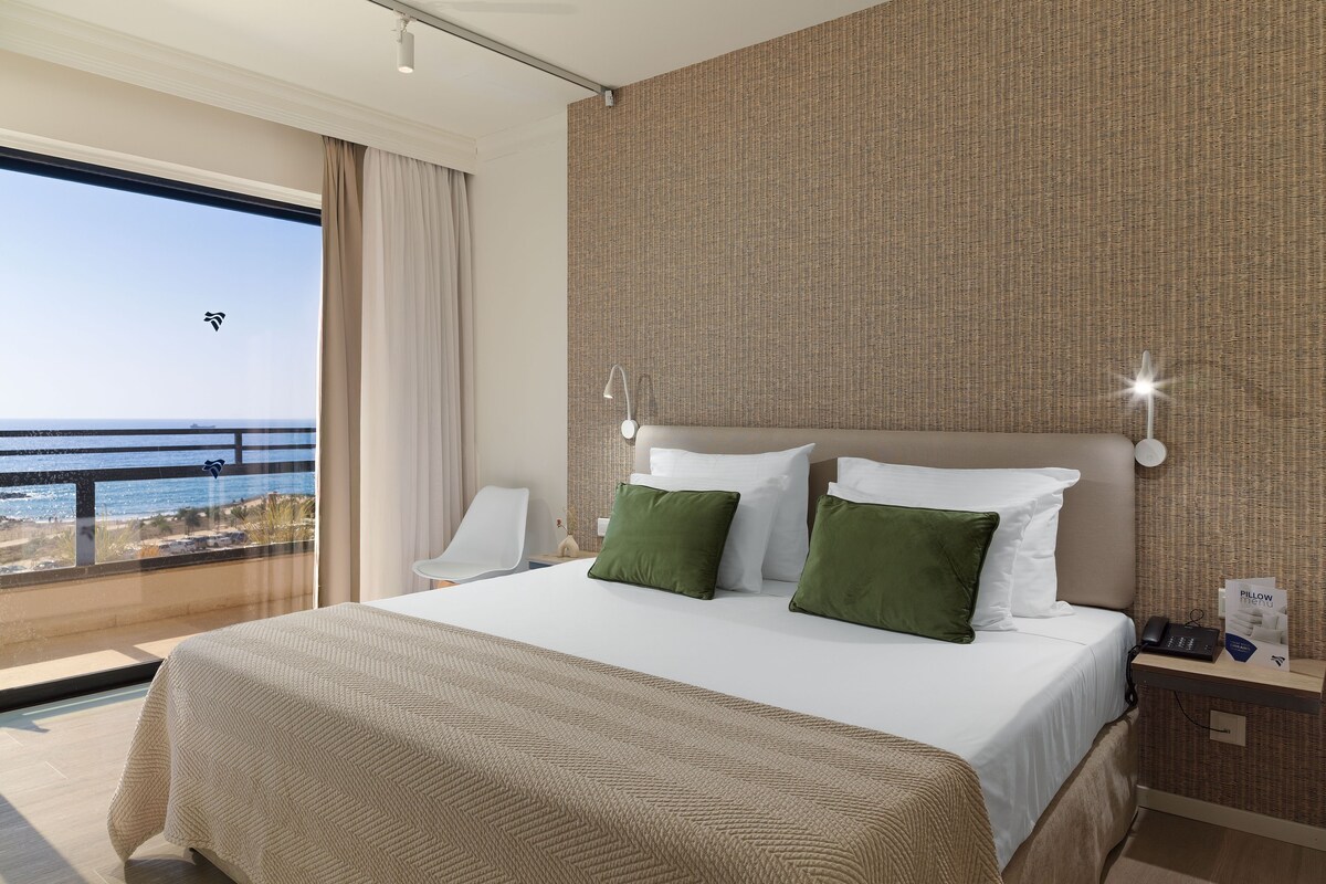 A hotel room with a large bed, two pillows, a nightstand, a lamp, and a view of the ocean.