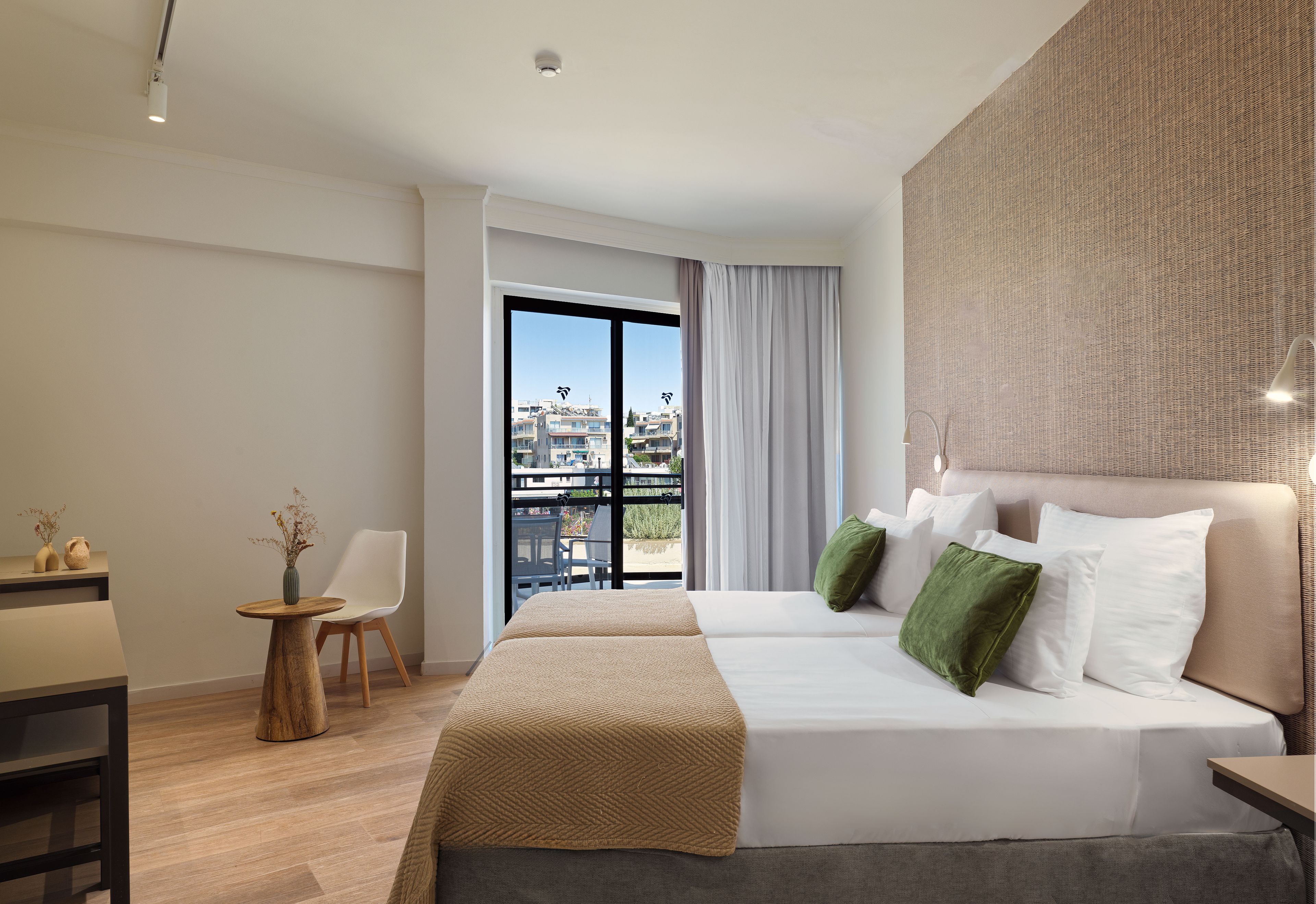 A hotel room with a balcony, a bed with pillows, a chair, a small table with a vase, and a wall with a textured pattern.