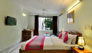Habitación de hotel con cama, mesita de noche, lámpara, televisor y ventana con vistas a edificios y árboles.
