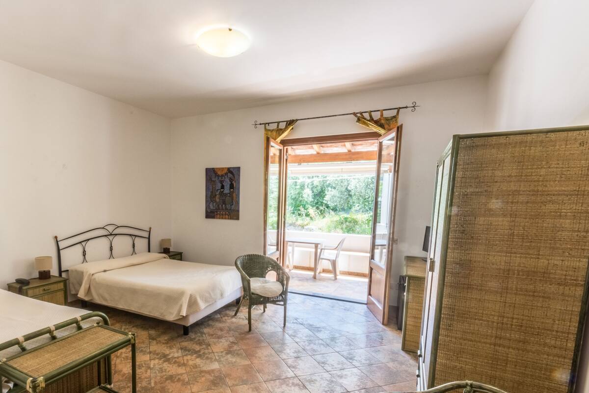A bedroom with a bed, a chair, a small table, and a balcony with a view of greenery.