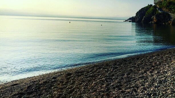 Une plage de galets avec une eau calme et une falaise rocheuse qui se dessine à l’horizon.