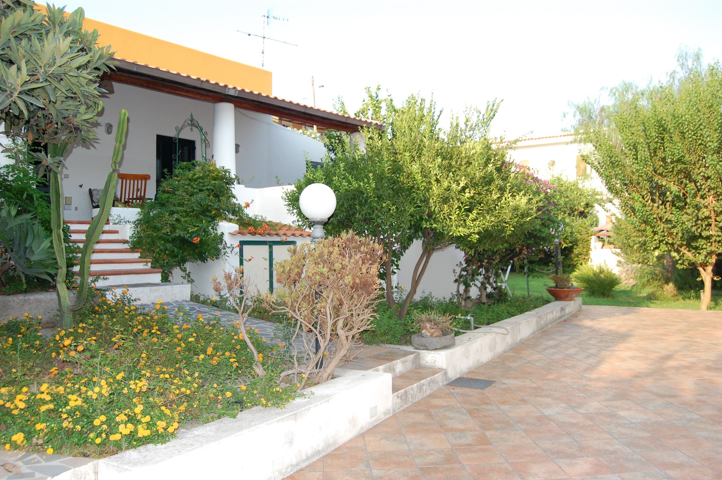 Una casa con un patio empedrado, un pequeño jardín con flores amarillas y un árbol.