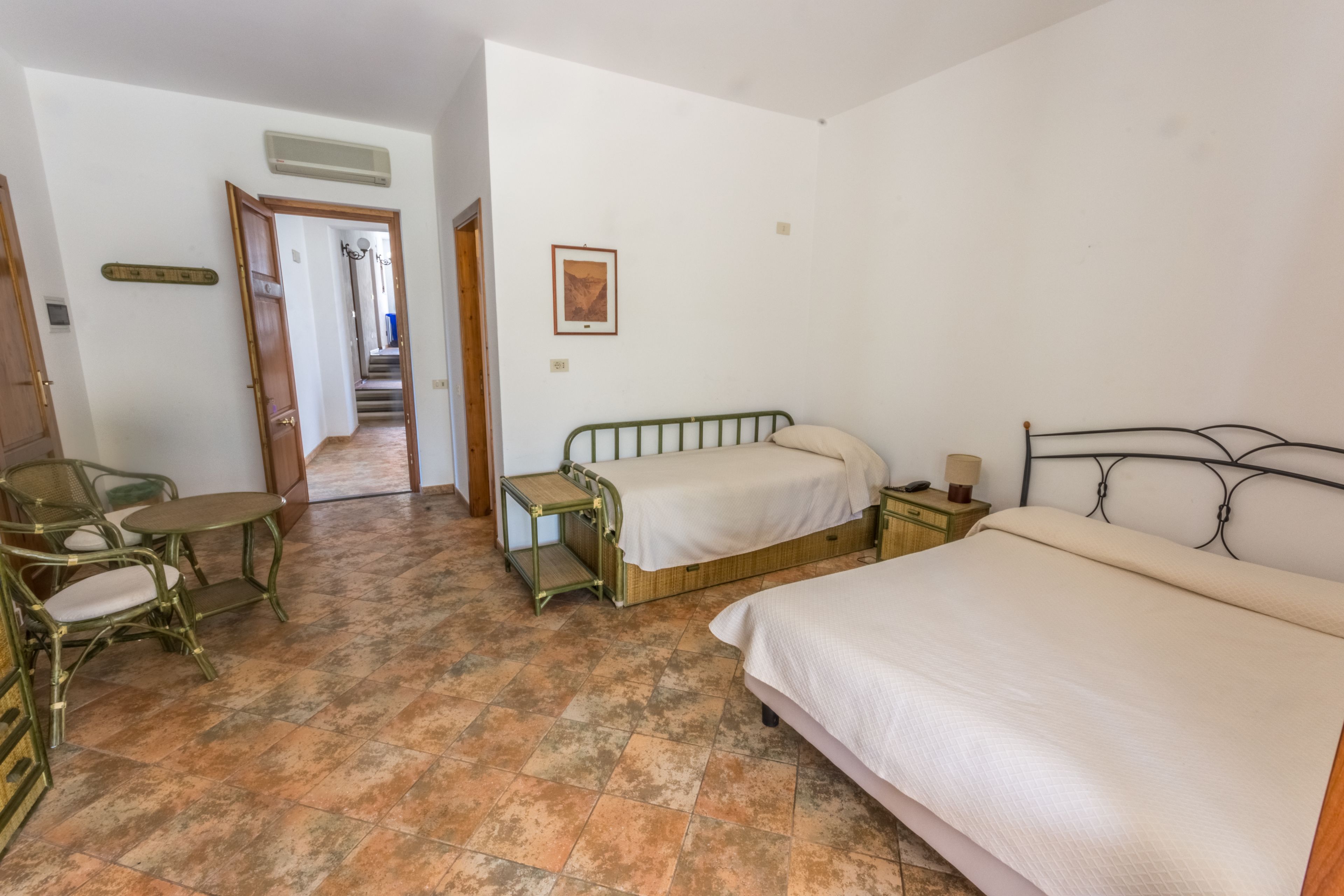 Un dormitorio con una cama grande, una mesita de noche, una silla y una pequeña mesa. Hay una puerta que da a un pasillo.