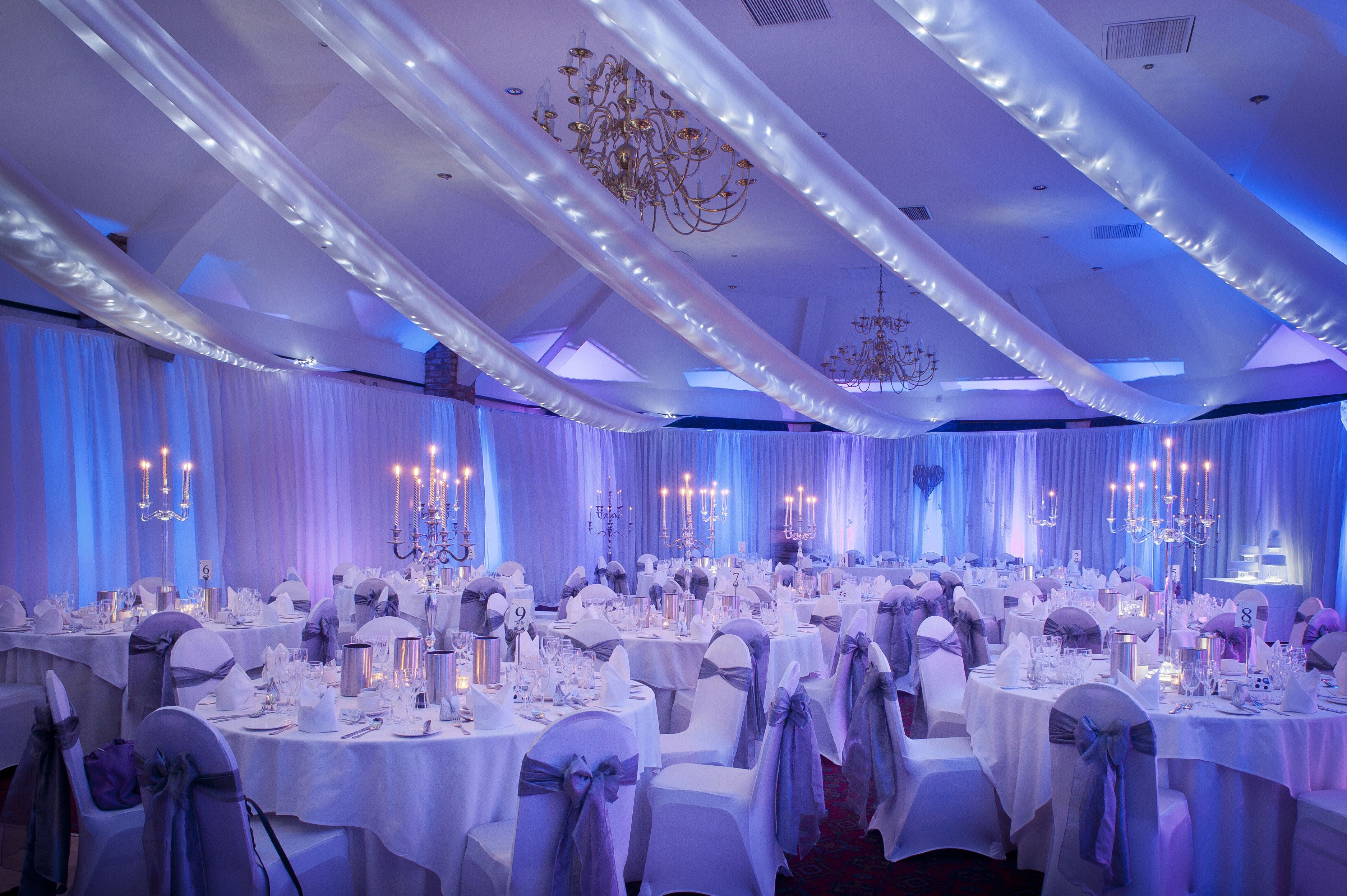 A banquet hall with round tables set for a formal event, featuring white tablecloths and chairs with blue sashes.
