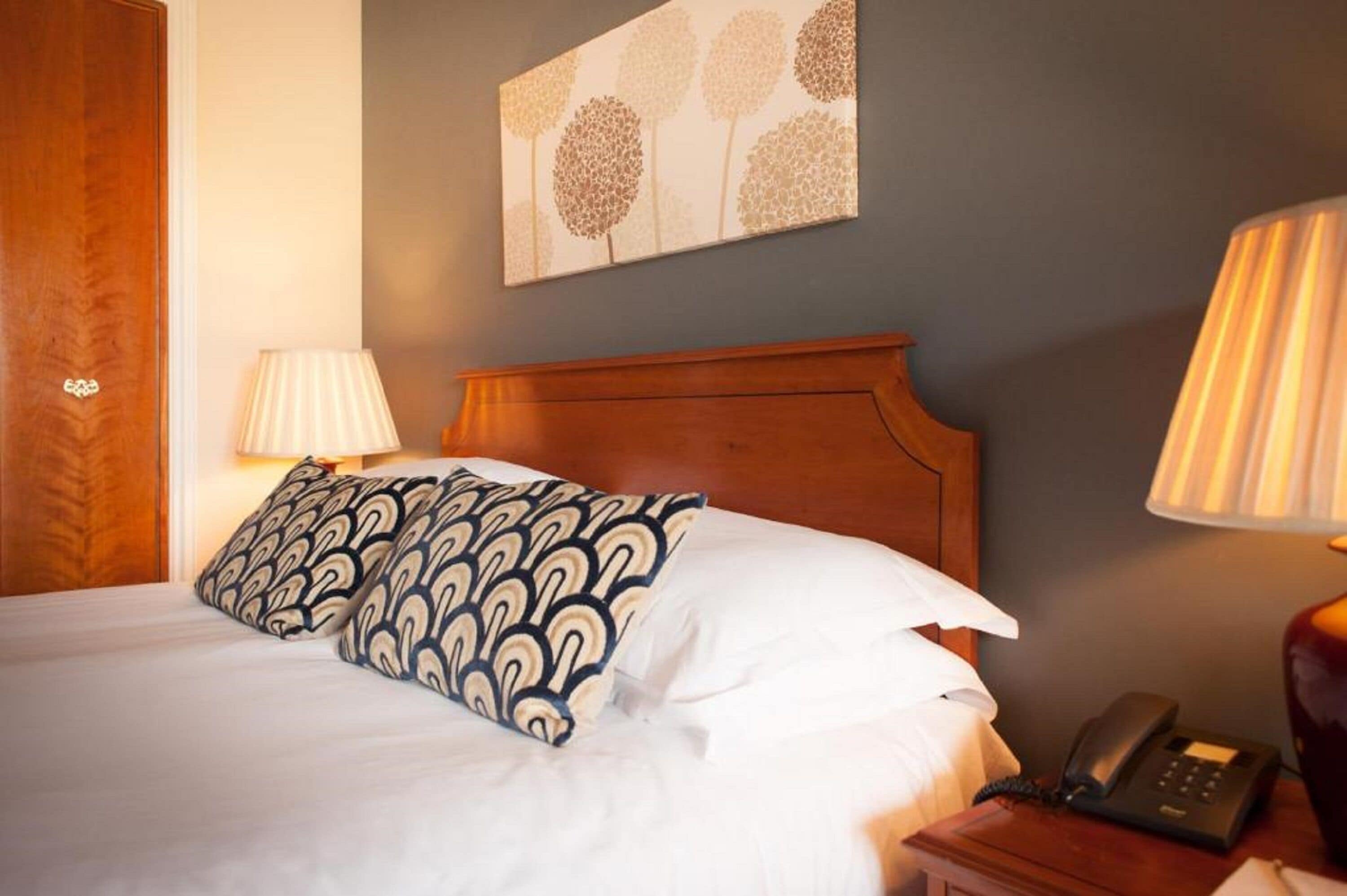 A neatly made bed with patterned pillows, a wooden headboard, a bedside lamp, and a telephone on a wooden nightstand.