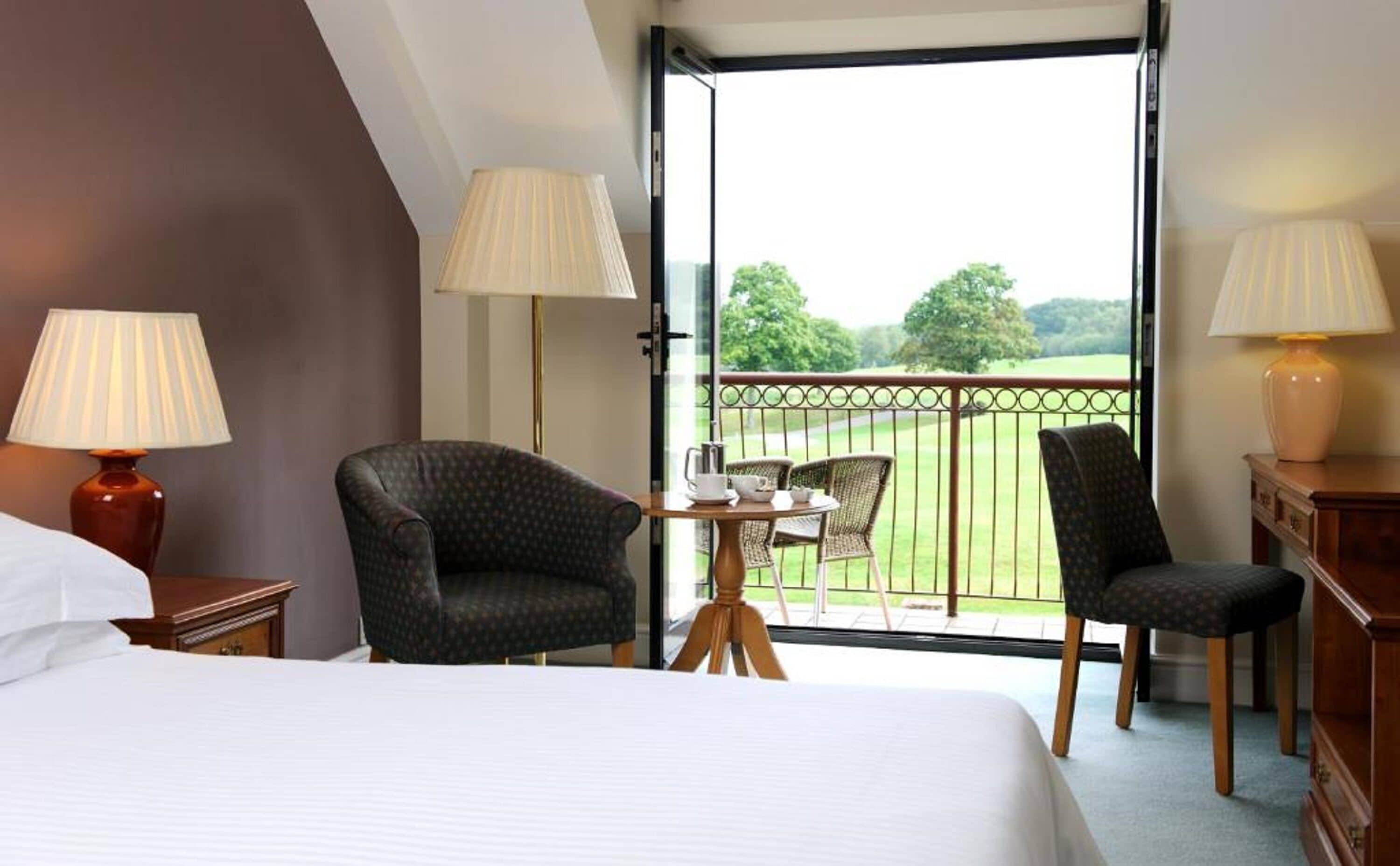 Habitación de hotel con cama, dos sillas, una mesita con taza y plato, y un balcón con vista a un paisaje verde.