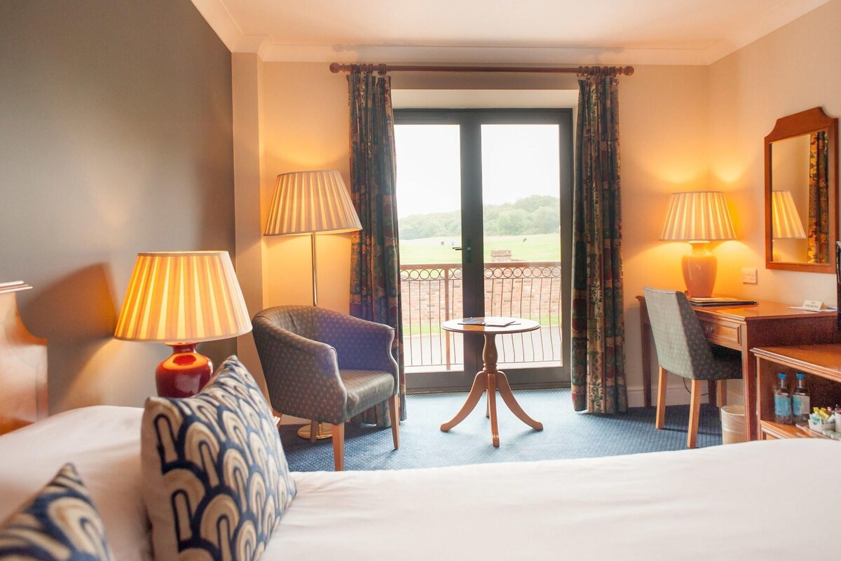 Habitación de hotel con cama, dos sillas, un escritorio, una mesita y dos lámparas. Tiene un balcón con vistas al exterior.