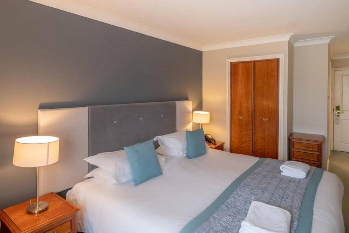 A hotel room with a bed, two bedside lamps, a wooden wardrobe, and a small bedside table.