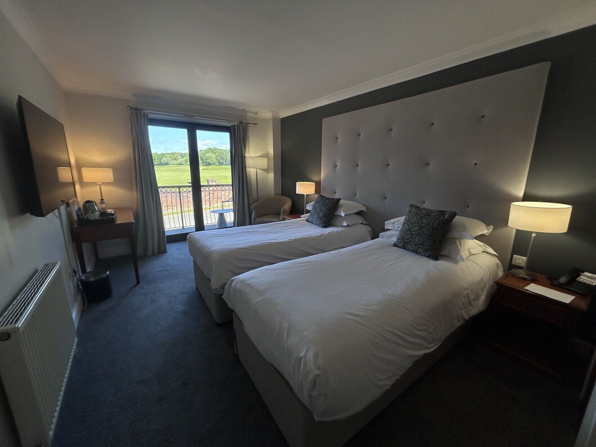 Habitación de hotel con dos camas, un escritorio, una silla, una lámpara y vistas a un paisaje verde.