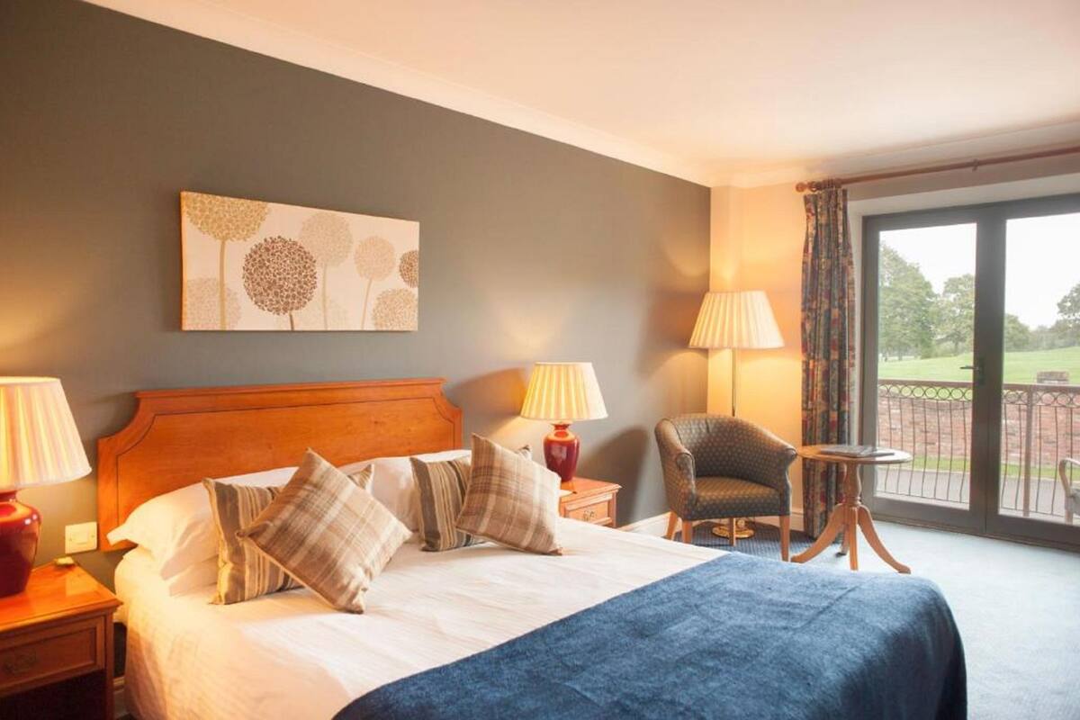 A hotel room with a bed, two bedside lamps, a chair, a small table, and a view of the outdoors through a large window.