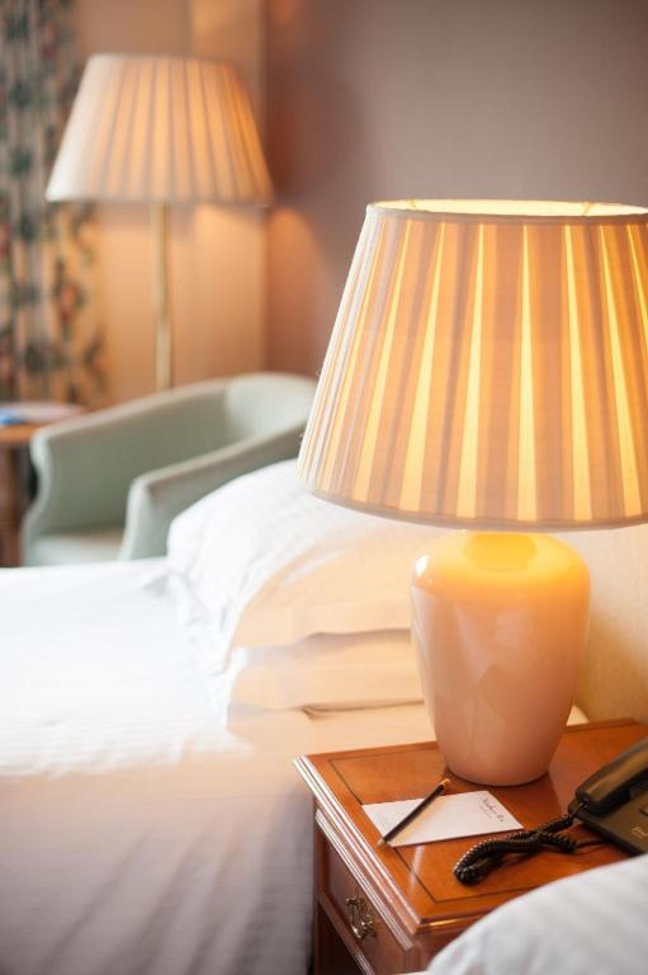 Een net opgemaakt bed met witte lakens, een bedlampje, een nachtkastje met een telefoon en notitieboekje, en een wandlamp op de achtergrond.