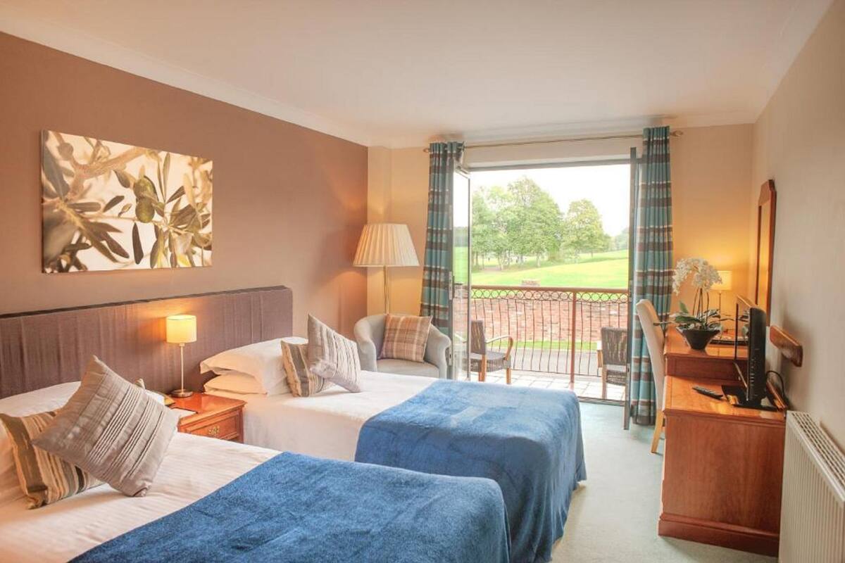 A hotel room with two beds, a desk, a chair, a lamp, and a view of a garden through a sliding door.
