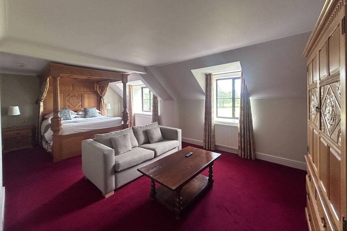 A bedroom with a four-poster bed, a sofa, a wooden coffee table, and a large window with curtains.