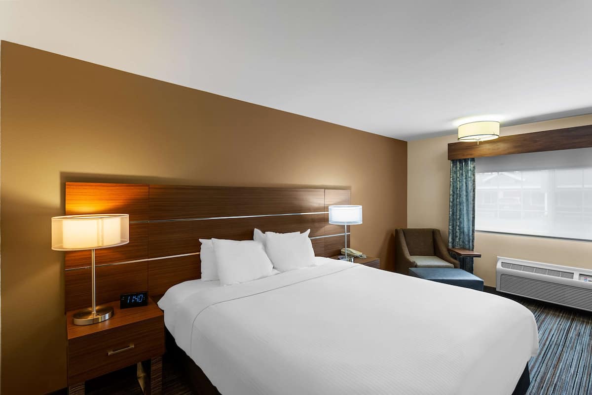 A hotel room with a large bed, a nightstand with a lamp and clock, a chair, and a window with curtains.