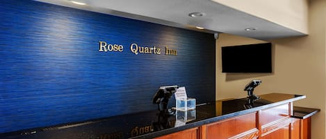 Een receptiebalie met een blauwe, gestructureerde wand en de naam 'Rose Quartz Inn' in gouden letters. Er zijn twee kaartlezers en een glazen vitrine.