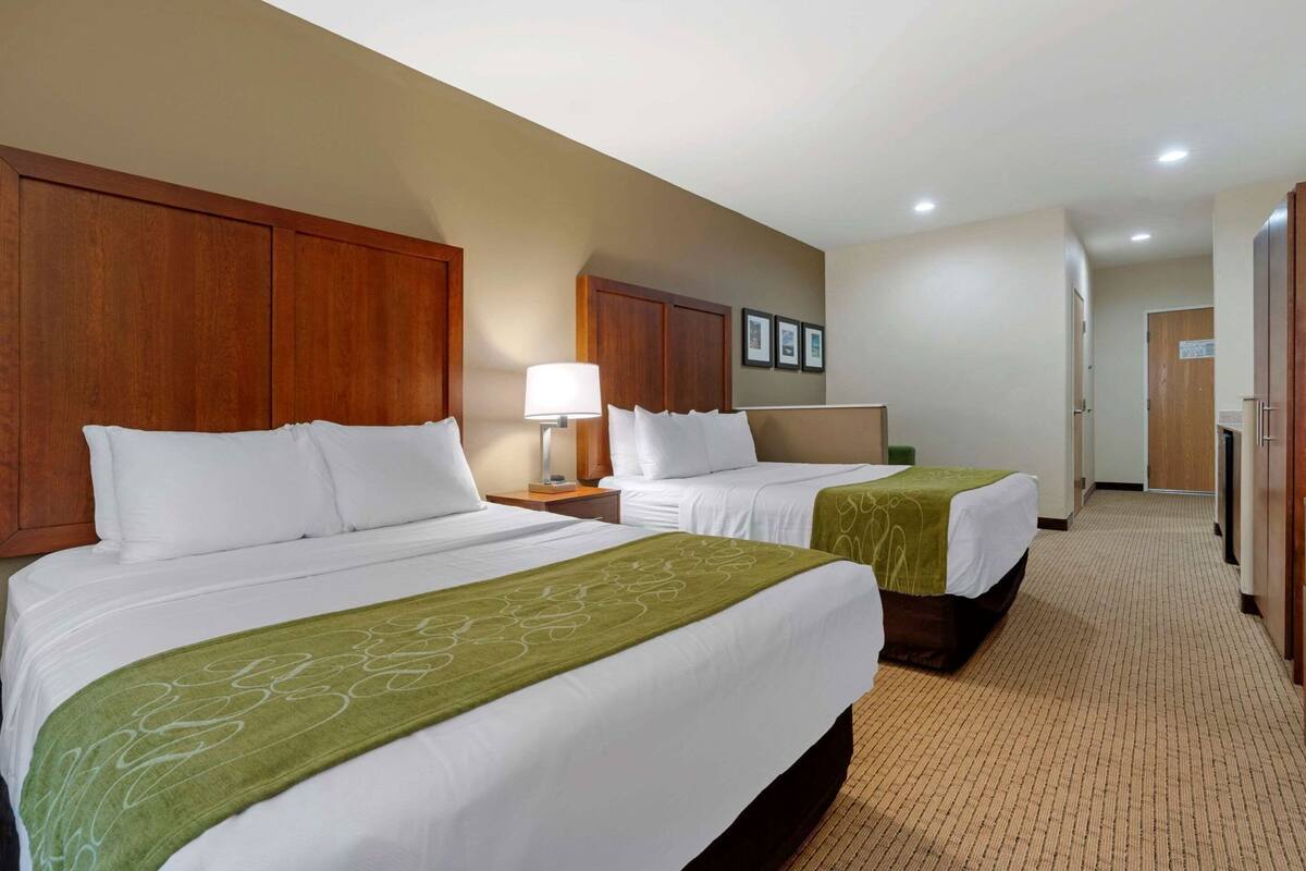 A hotel room with two beds, a nightstand with a lamp, a wooden wardrobe, and a door leading to a hallway.