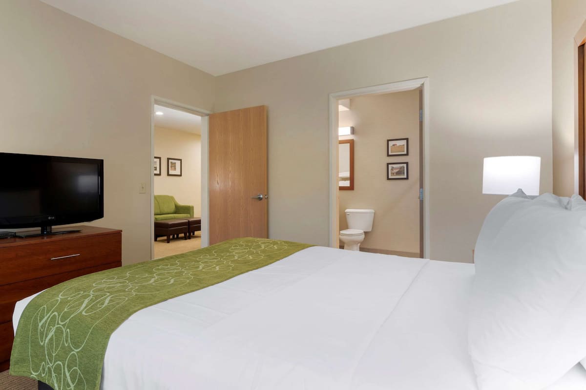 A hotel room with a bed, a TV on a wooden dresser, a green throw blanket, a white pillow, and a door leading to a bathroom.