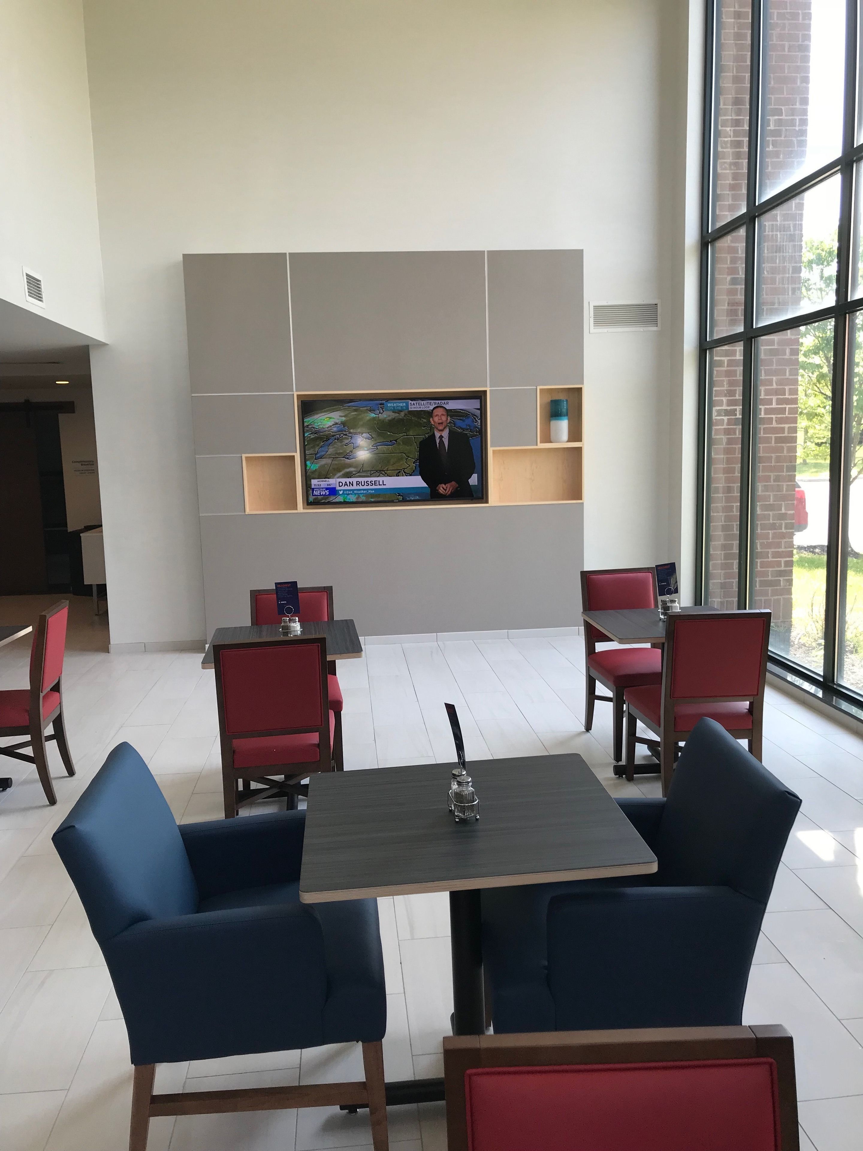 Um interior moderno com uma televisão exibindo um noticiário, arranjos de assentos e vista através de grandes janelas.