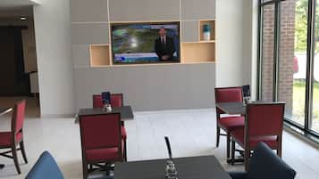 Um interior moderno com uma televisão exibindo um noticiário, arranjos de assentos e vista através de grandes janelas.