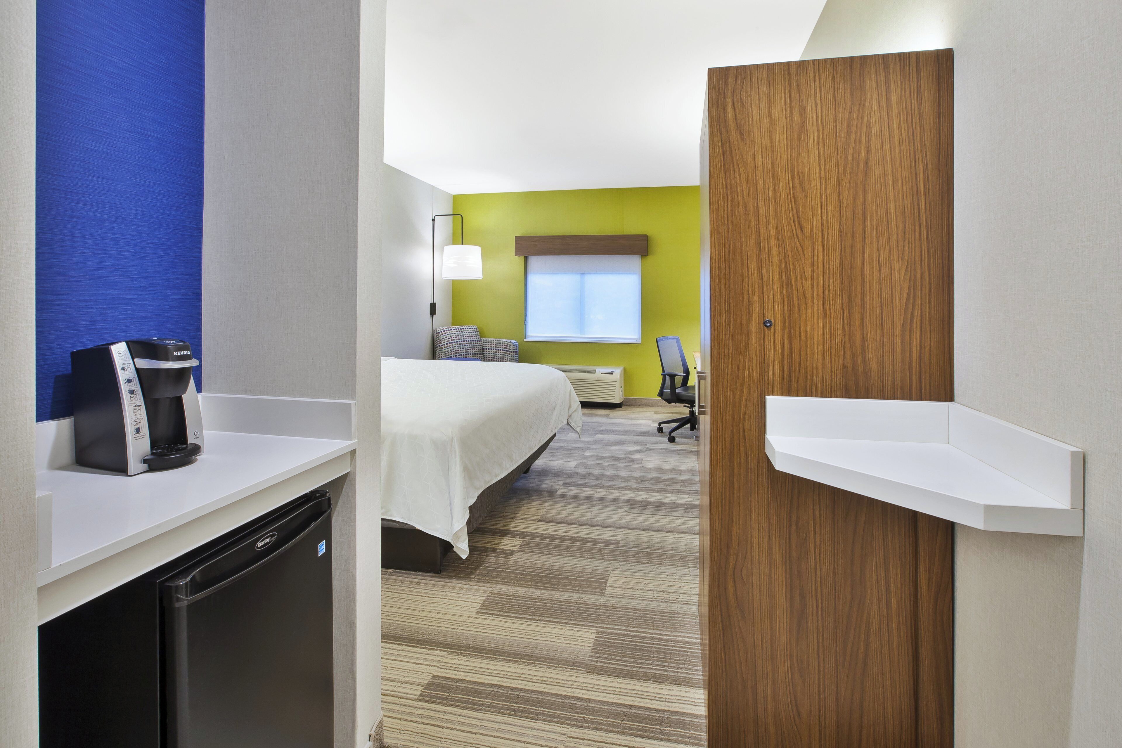 A hotel room with a bed, a desk with a coffee maker, a chair, and a window with blinds.