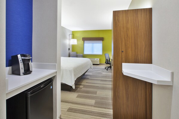 A hotel room with a bed, a desk with a coffee maker, a chair, and a window with blinds.