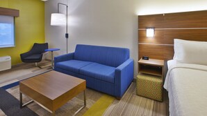 A hotel room with a blue sofa, a wooden coffee table, a white bed, and a small wooden nightstand with a lamp. - Holiday Inn Express Hotel & Suites Rochester - Victor by IHG (Victor)