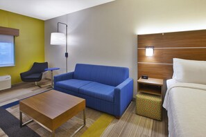 A hotel room with a blue sofa, a wooden coffee table, a white bed, and a small wooden nightstand with a lamp. - Holiday Inn Express Hotel & Suites Rochester - Victor by IHG (Victor)