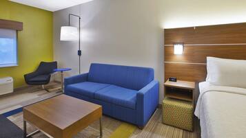 A hotel room with a blue sofa, a wooden coffee table, a small side table, a bed, and a nightstand with a clock.