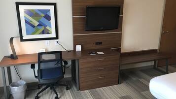 A hotel room with a wooden desk, a chair, a television mounted on the wall, a bed, and a framed abstract artwork.