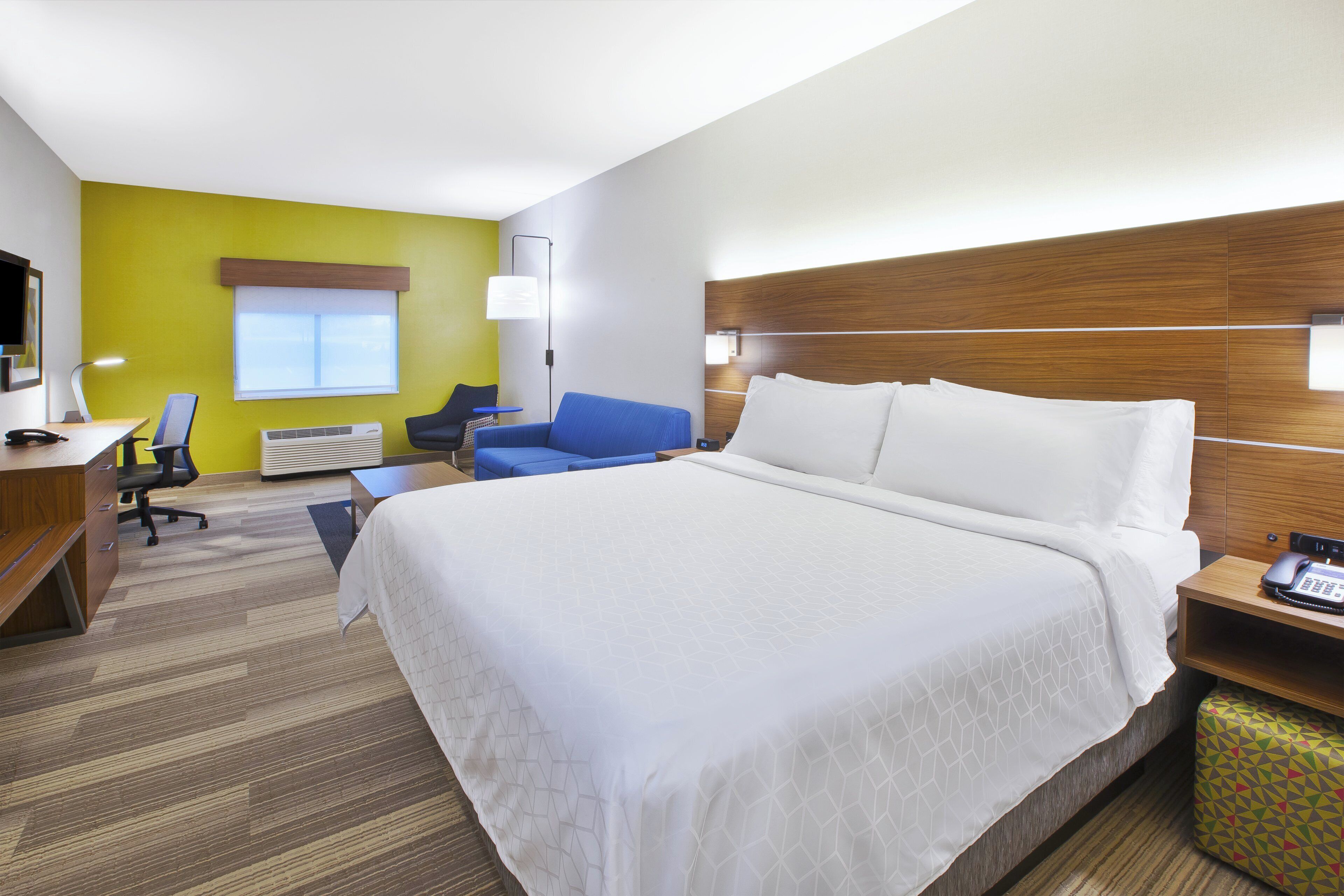 Quarto de hotel com uma cama grande, uma escrivaninha, uma cadeira, uma mesinha e uma mesa de cabeceira com abajur.