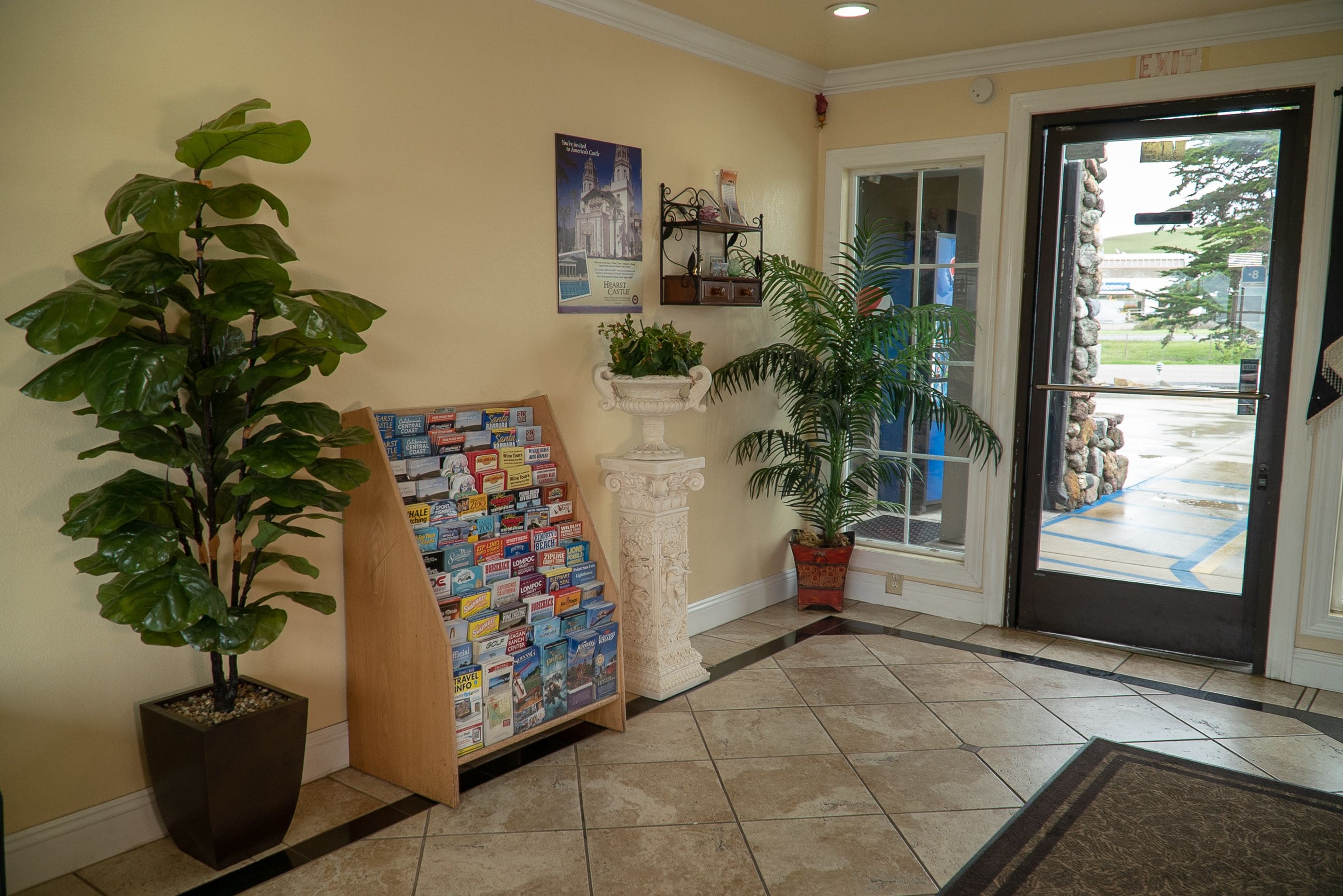 A hotel lobby with a large potted plant, a travel information board, and a door leading outside.