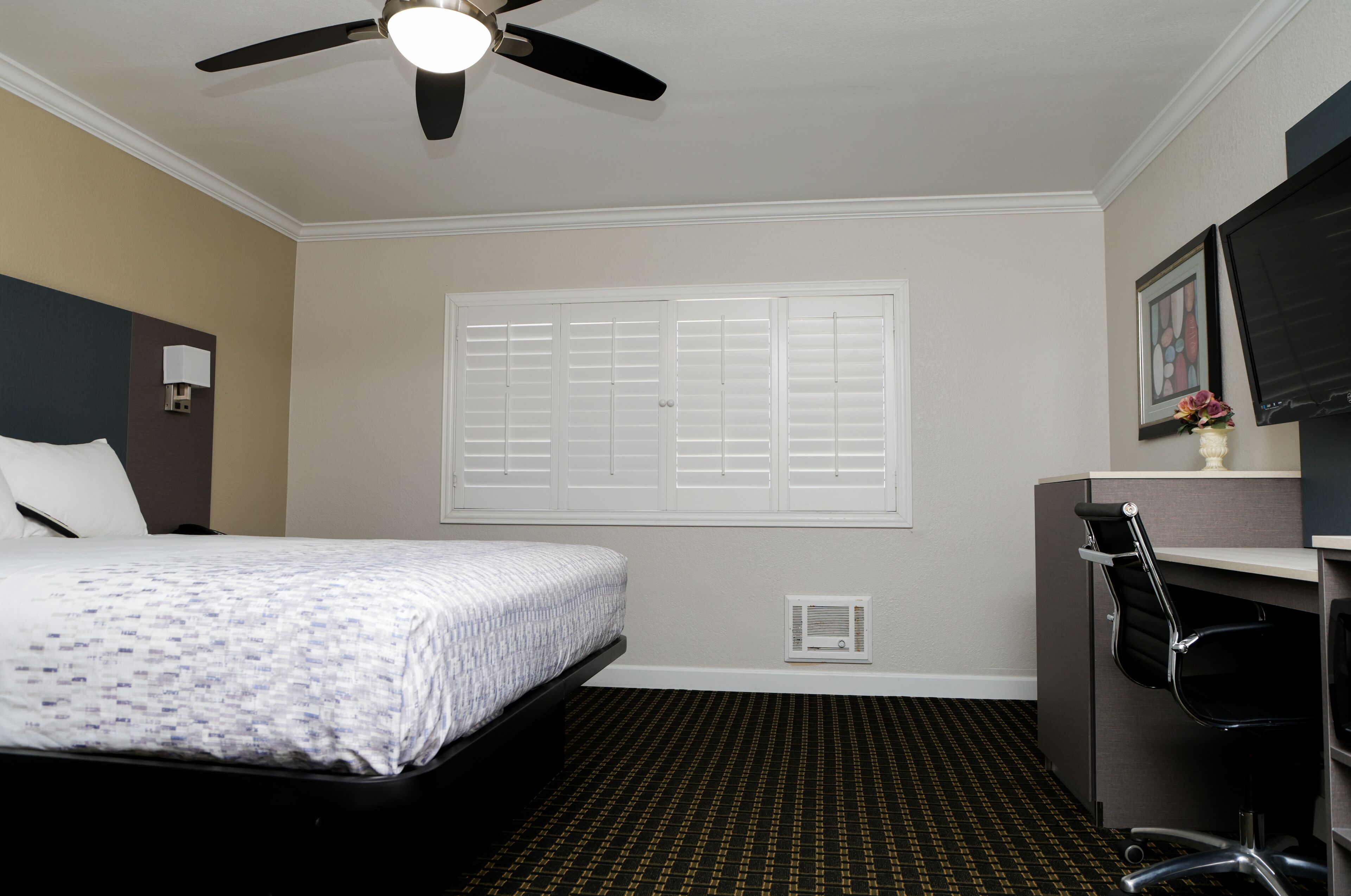 Habitación de hotel con cama, escritorio con silla, ventilador de techo, ventana con persianas y televisor montado en la pared.