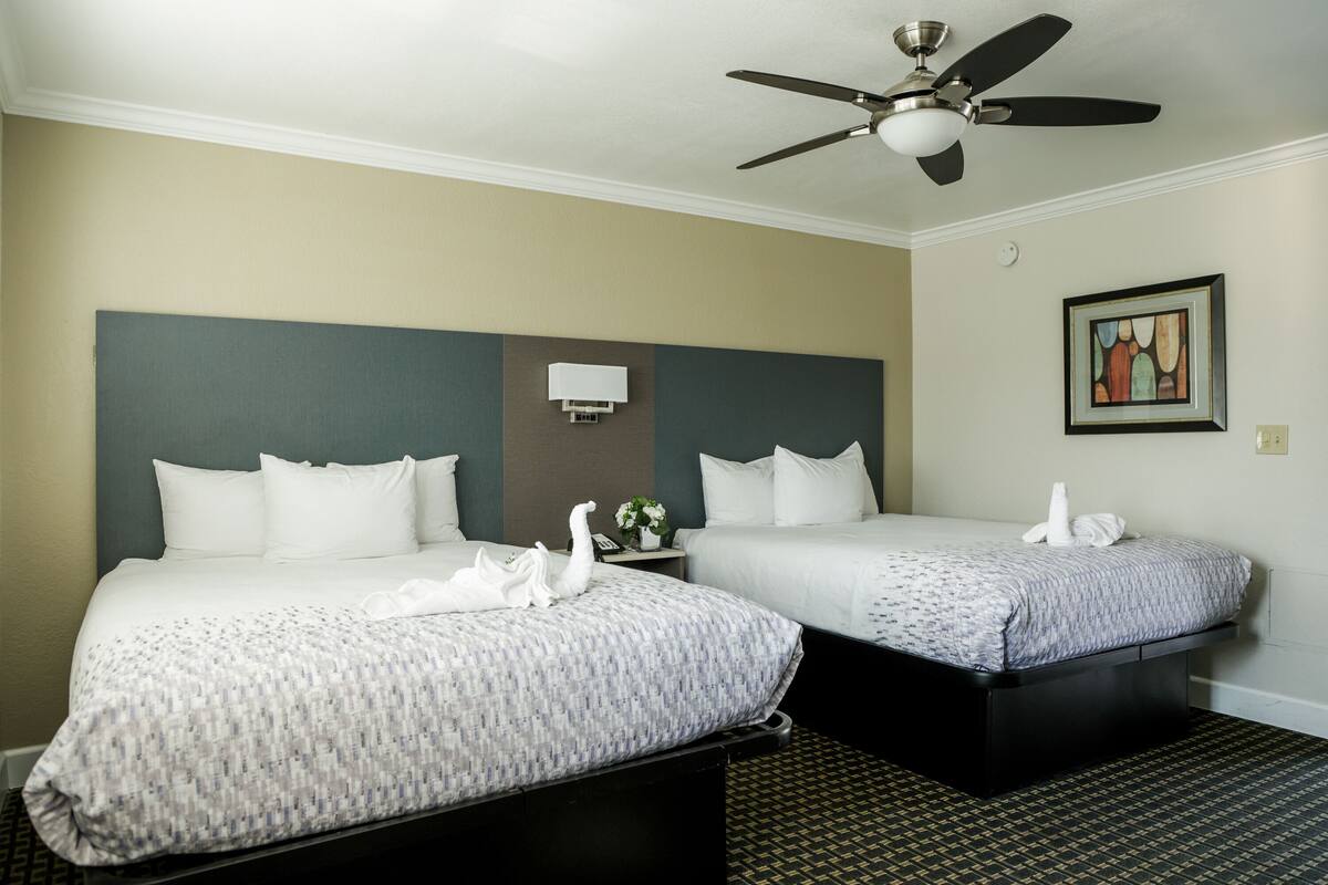 Habitación de hotel con dos camas, ventilador de techo, lámpara de pared y un cuadro enmarcado.