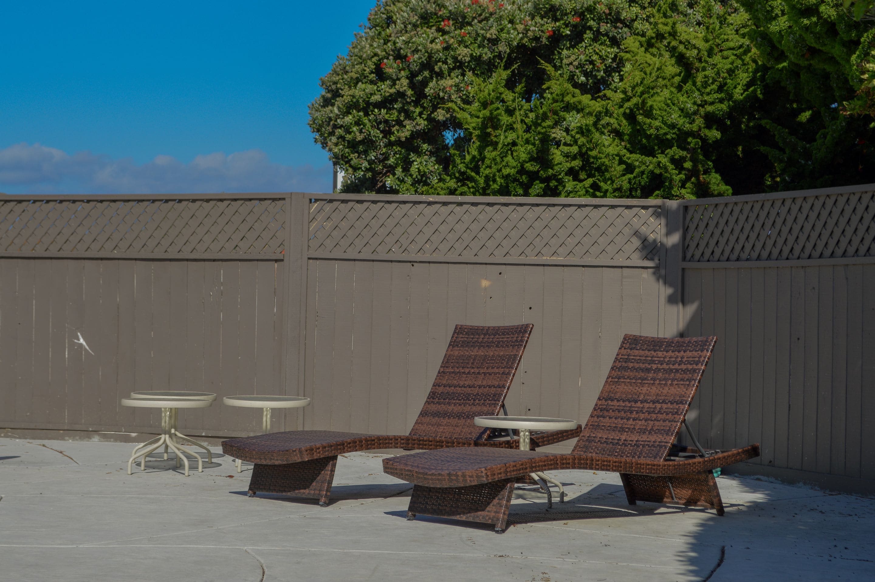 A sun lounge area with two lounge chairs and two small tables, set against a wooden fence and a large tree.