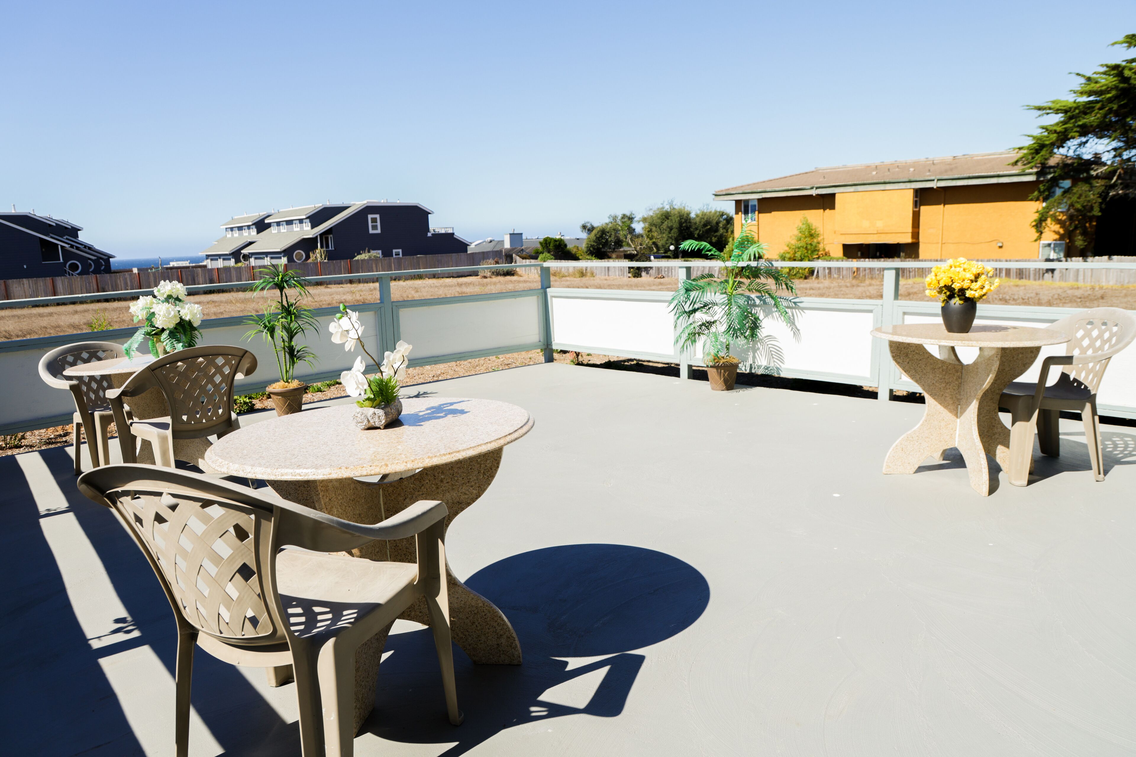 An outdoor patio area with round tables and chairs, potted plants, and a view of the ocean and residential buildings.