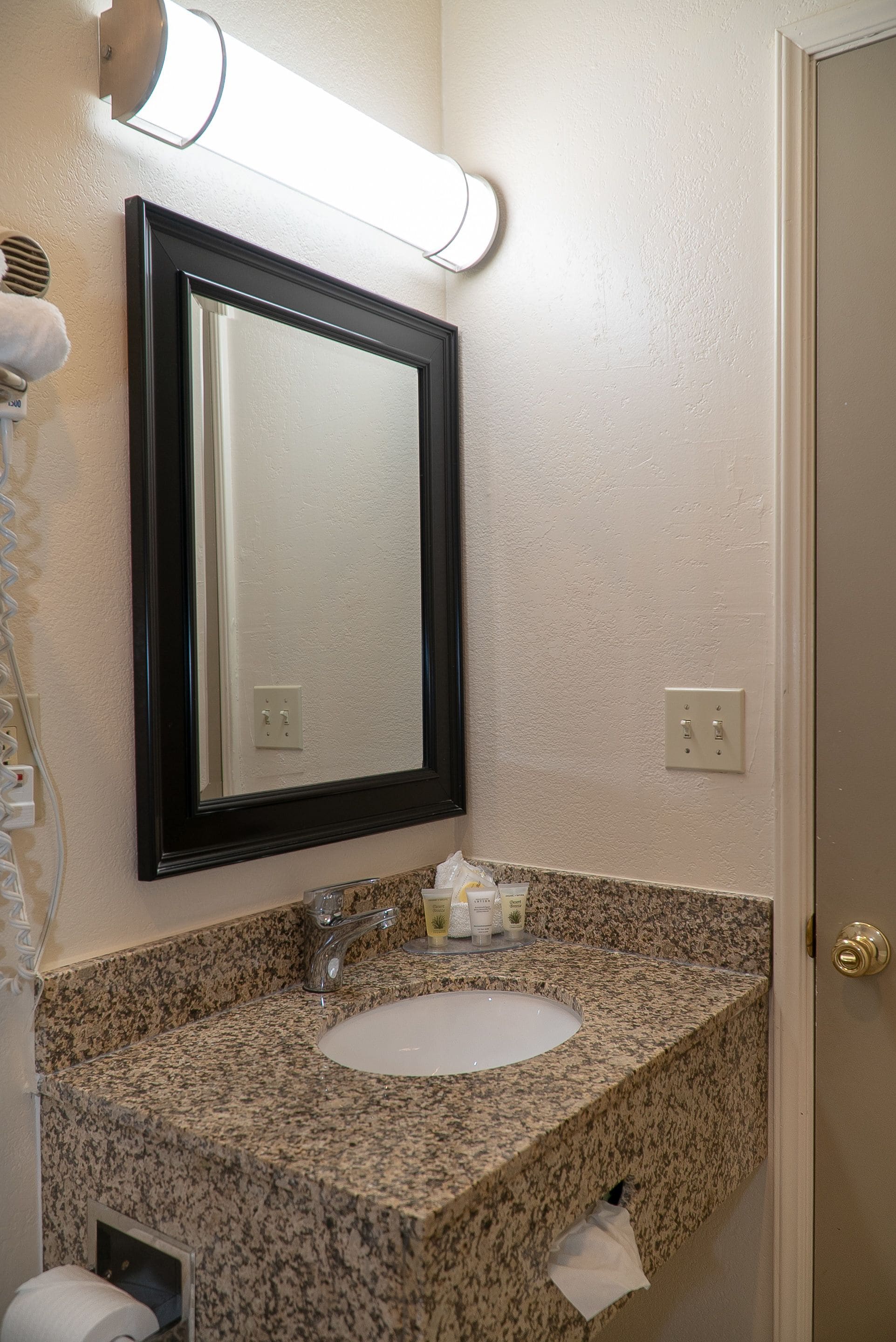 Baño con encimera de granito, lavamanos, espejo y dispensador de toallas.