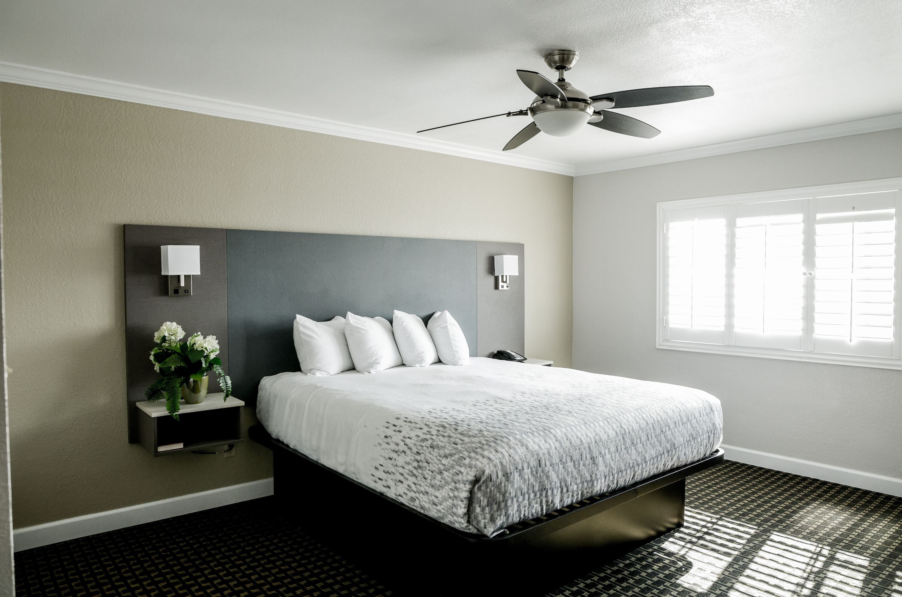 A hotel room with a large bed, a ceiling fan, a nightstand with a lamp, and a window with blinds.