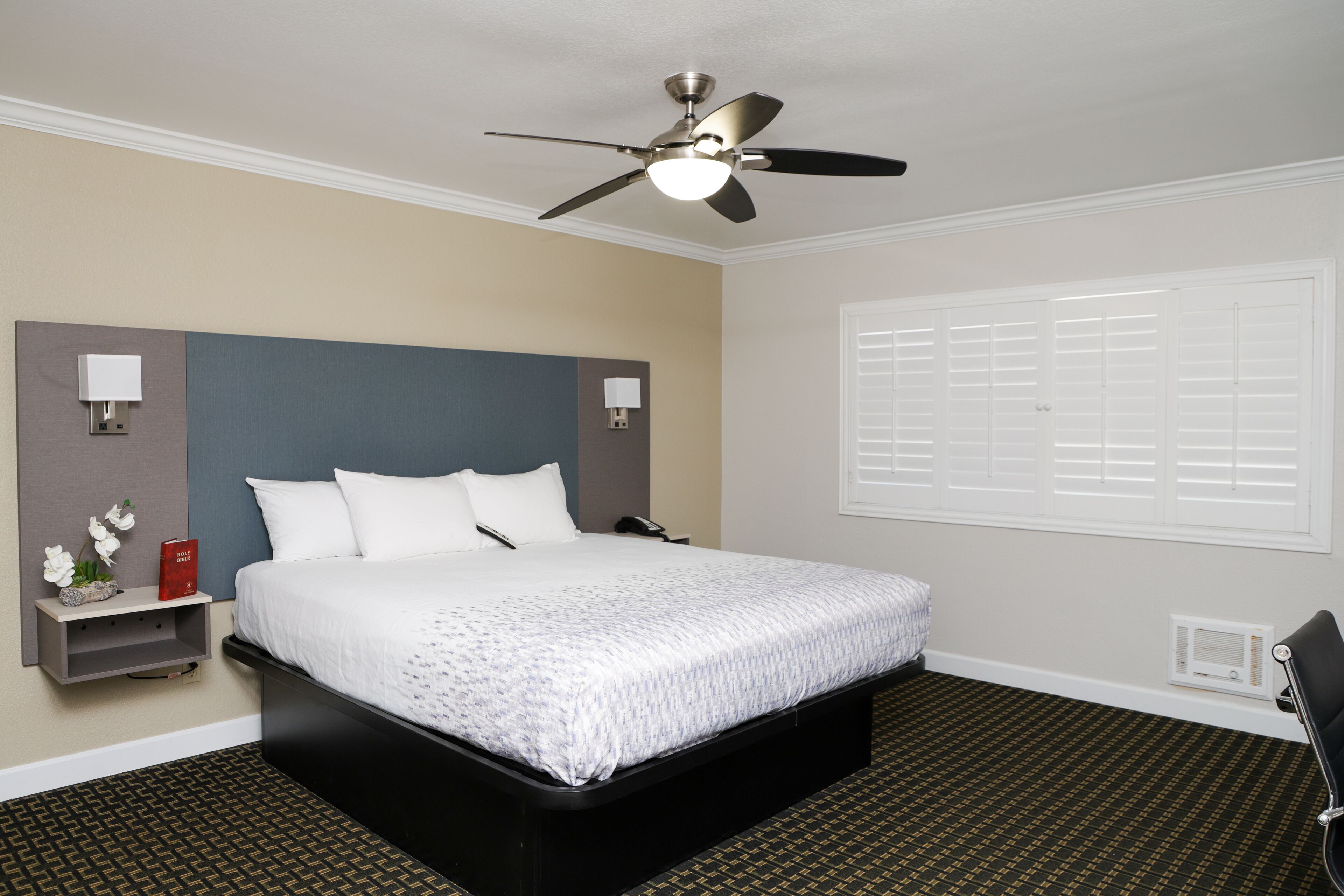 A hotel room with a large bed, a ceiling fan, a nightstand with a lamp, and a window with blinds.