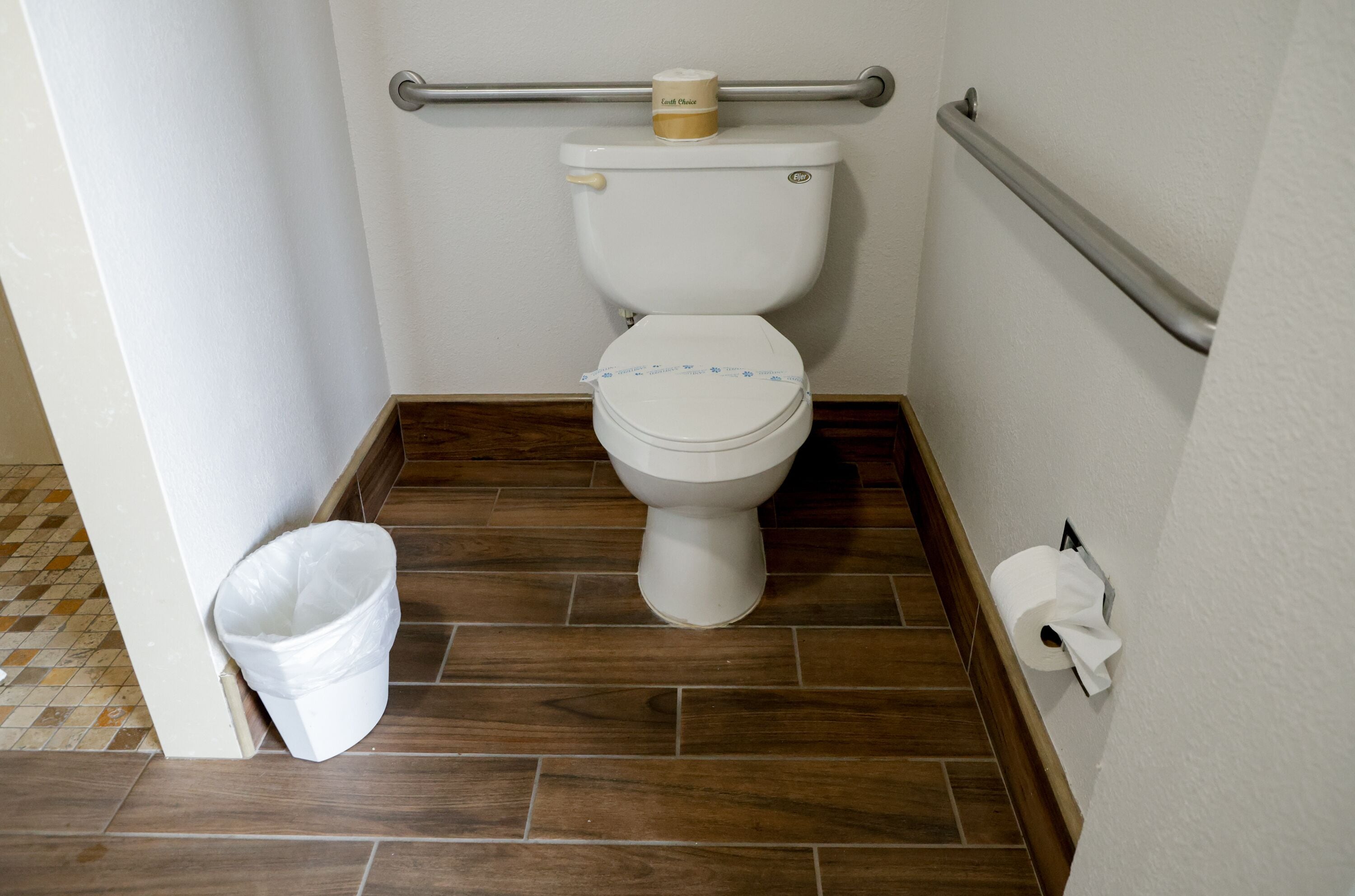A bathroom with a toilet, a handrail, a roll of toilet paper, and a trash bin.