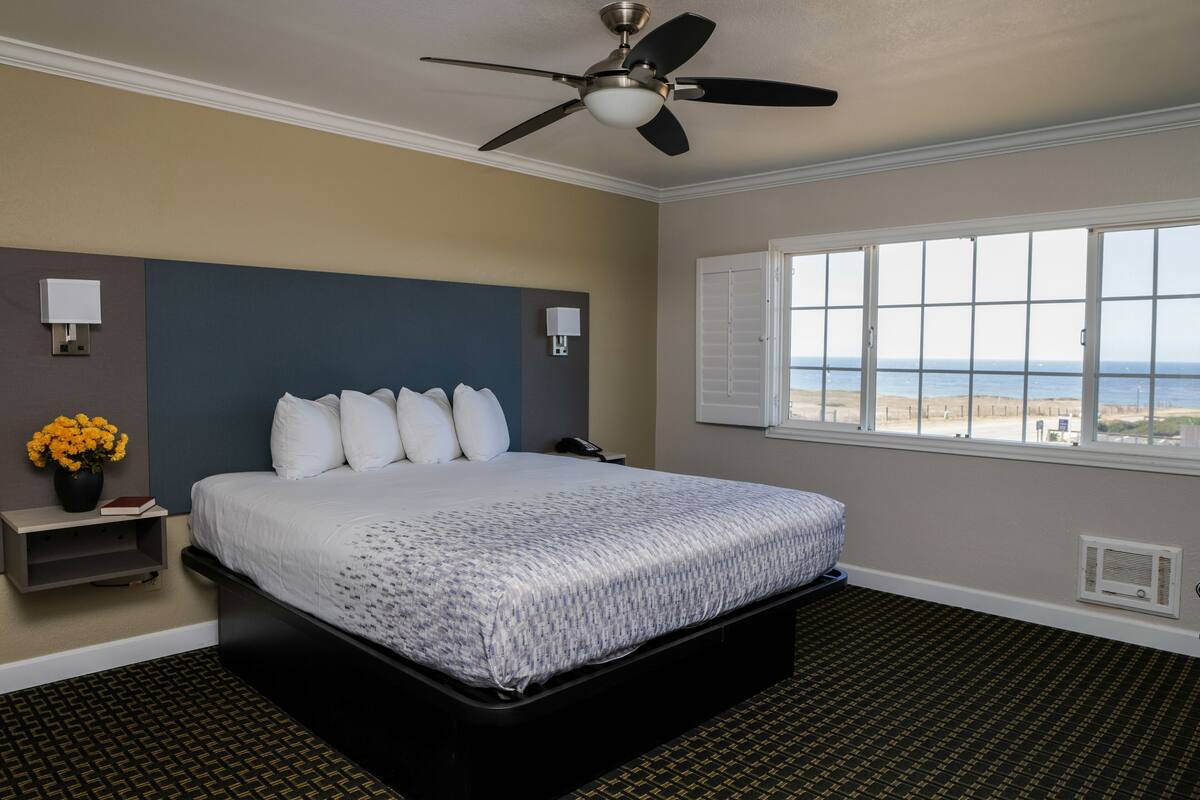 Habitación de hotel con una cama grande, un ventilador de techo, una mesita de noche con lámpara, una ventana con vistas a la playa y un pequeño estante con un florero.