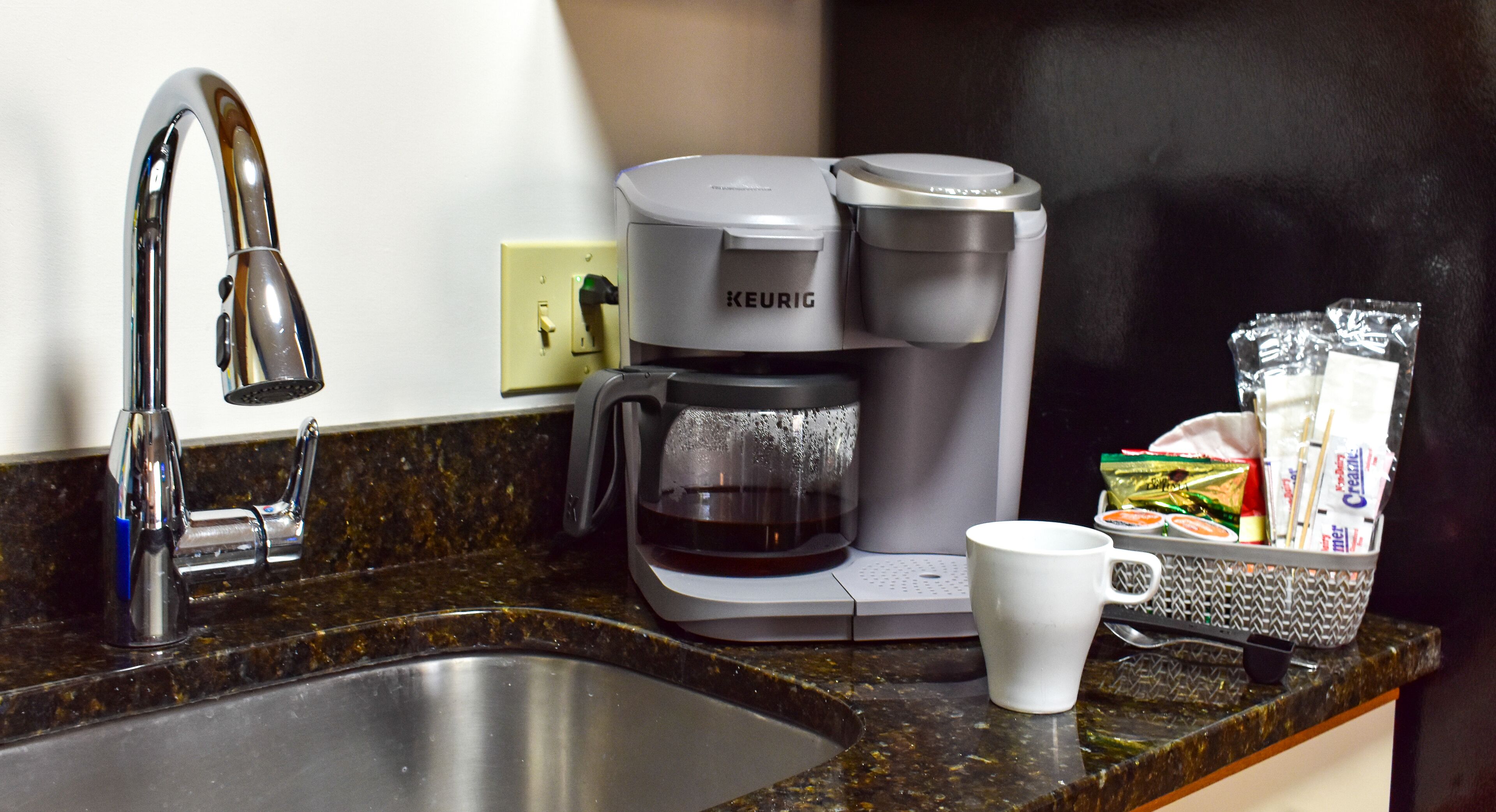 A kitchenette with a coffee maker, a sink, and a basket of condiments.