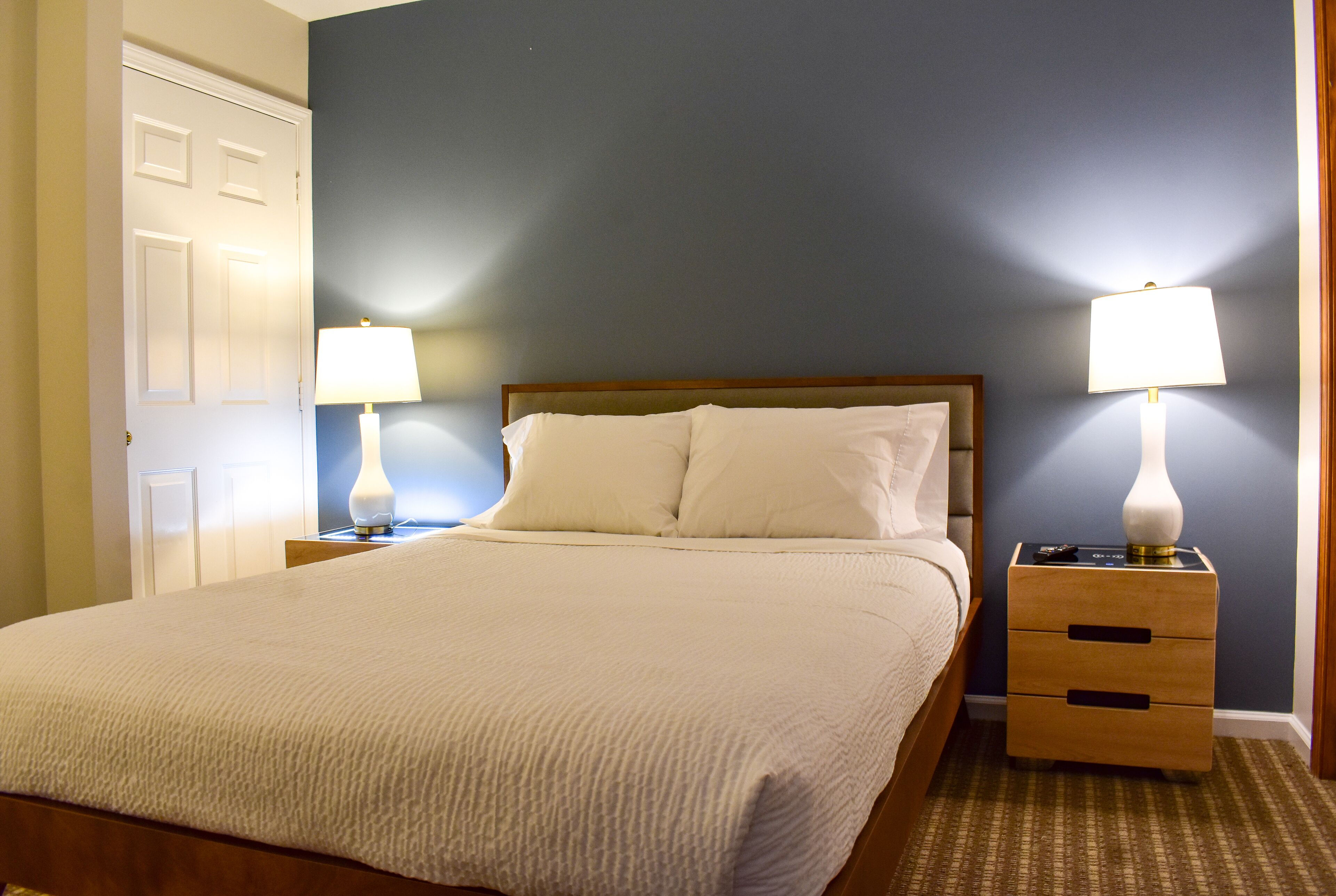 A neatly made bed with white linens, two bedside tables with lamps, and a door to the left.