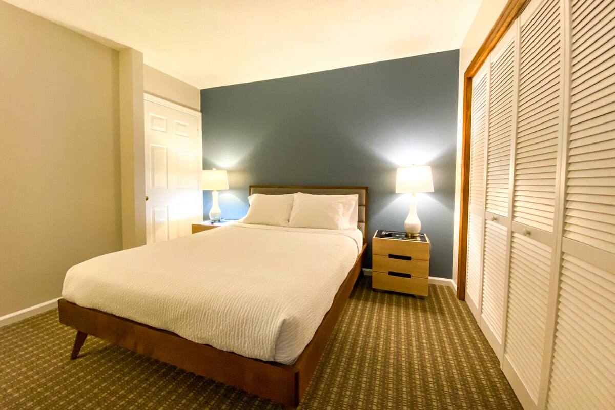 A neatly made bed with a wooden frame and white bedding, two bedside tables with lamps, a wardrobe with louvered doors, and a carpeted floor.