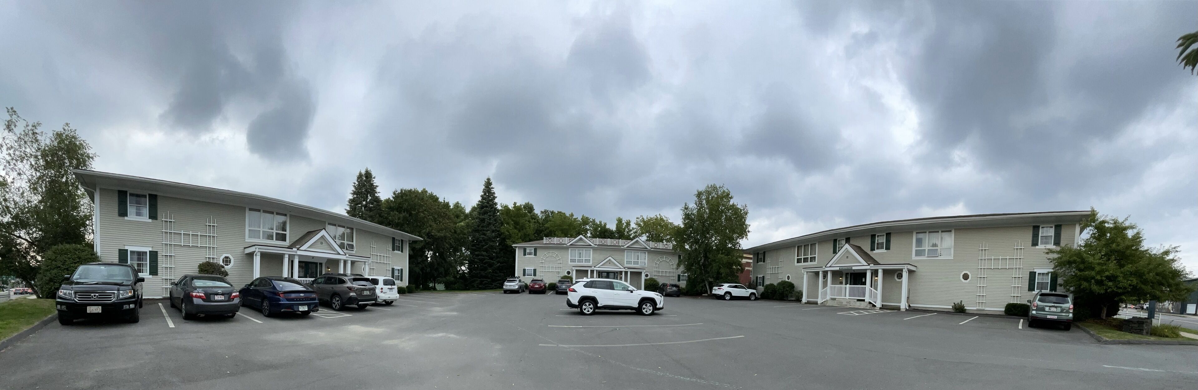 Un parking où plusieurs voitures sont garées devant un bâtiment à deux étages, entouré d’arbres.