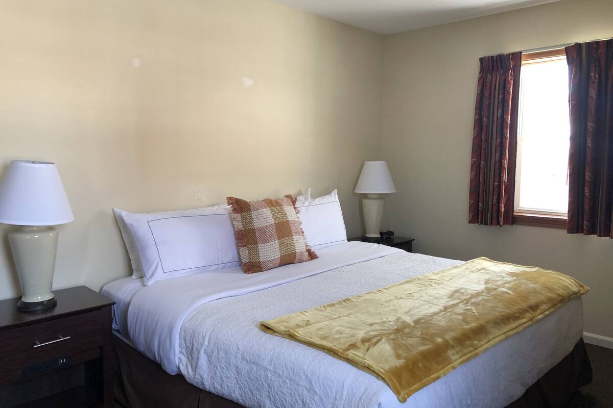 A neatly made bed with white bedding and a brown checkered throw, two bedside lamps, and a window with curtains.