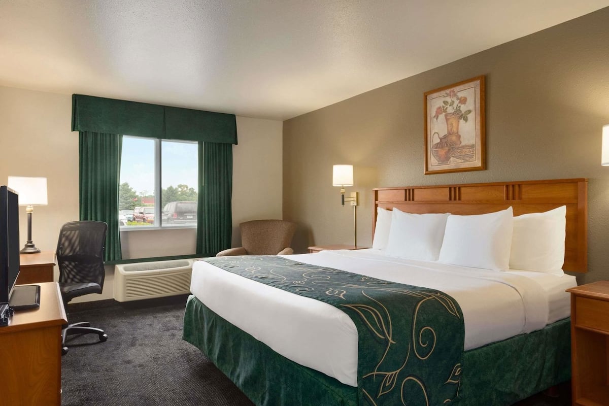 Una habitación de hotel con cama, escritorio con silla, ventana con cortinas, lámpara de pared y un cuadro.