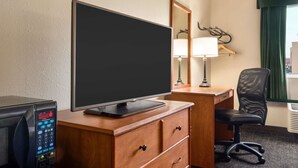 A hotel room with a wooden desk, a flat-screen TV, a microwave, a chair, a lamp, and a window with a view.