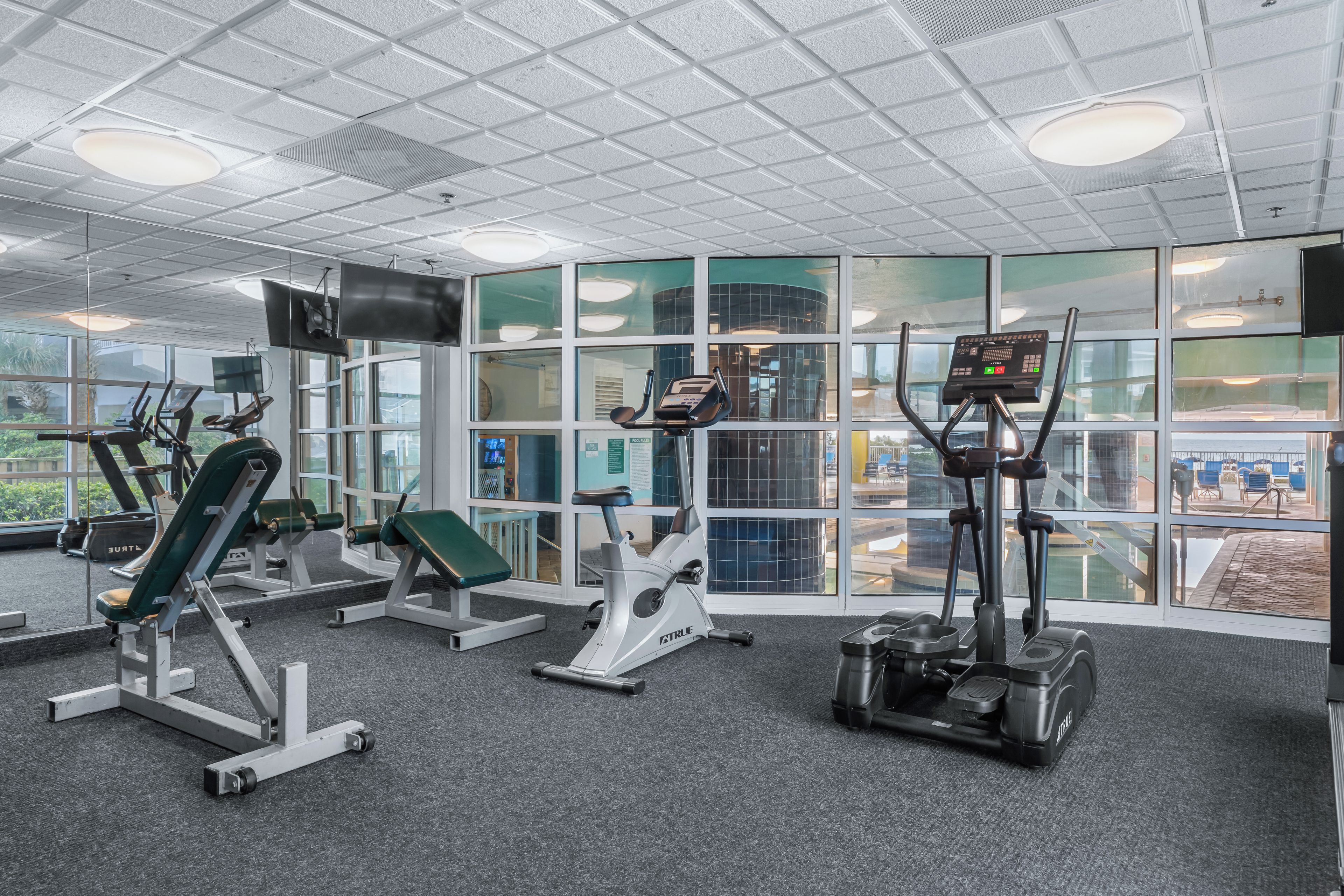 Une salle de sport équipée de divers appareils d’exercice, dotée de grandes fenêtres et d’un plafond orné d’un motif en réseau.